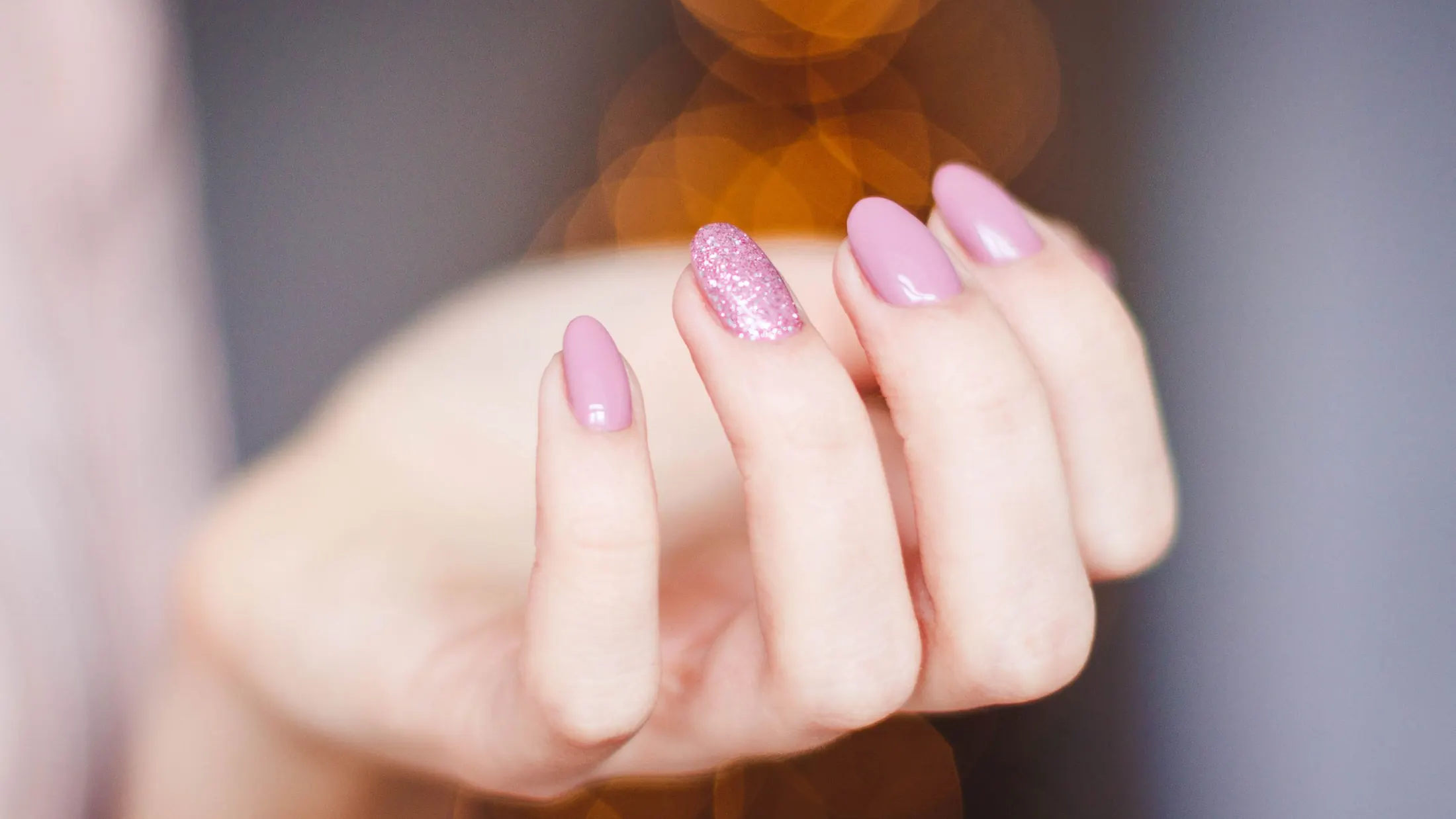 Main aux ongles roses, un ongle pailleté, sur fond de lumières dorées