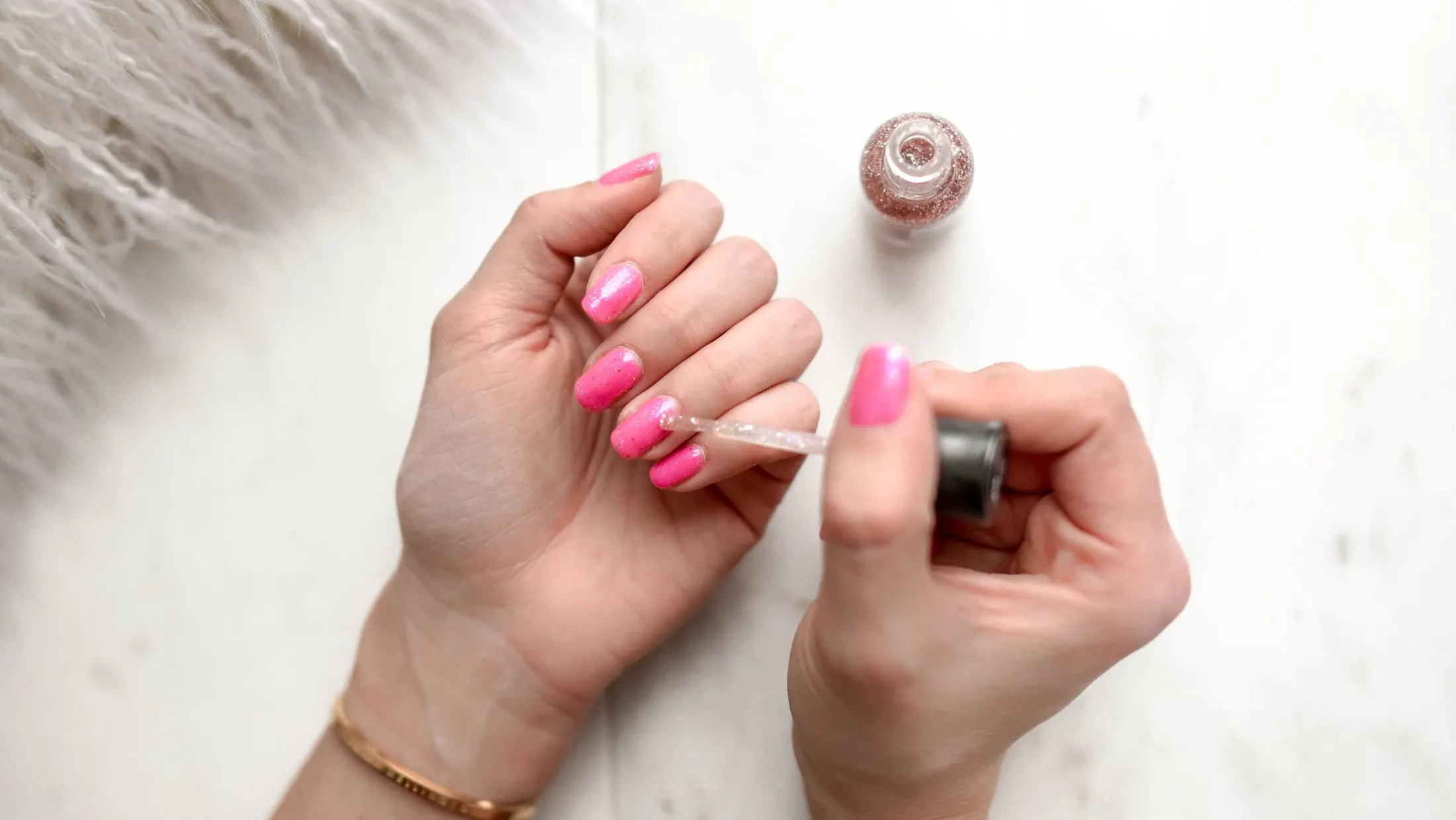 Mains vernies de rose, un flacon de vernis à paillettes posé sur du marbre